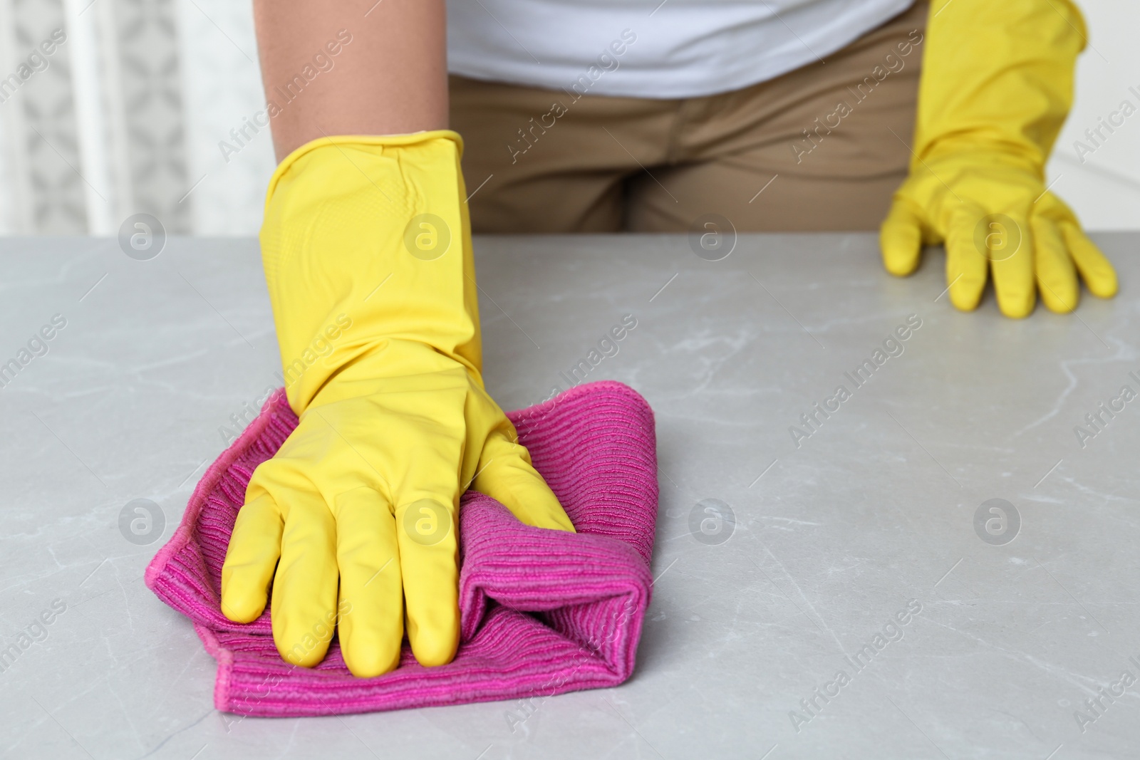 Photo of Woman in gloves wiping grey table with rag indoors, closeup