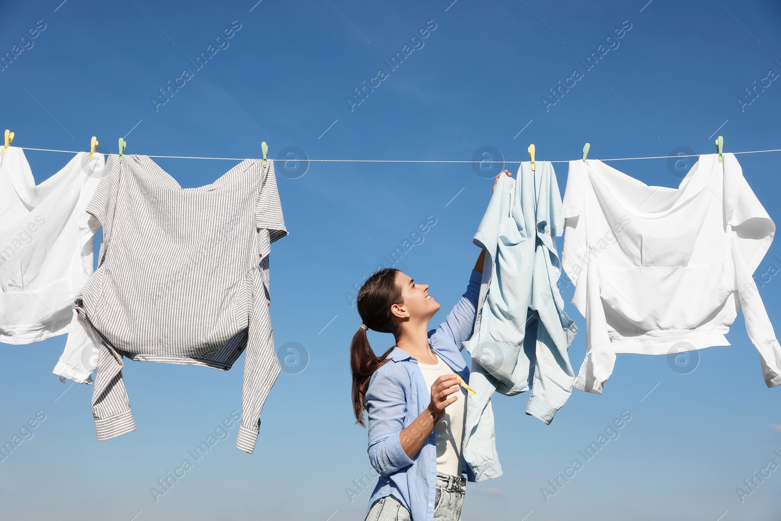 Photo of Woman hanging clothes with clothespins on washing line for drying against blue sky