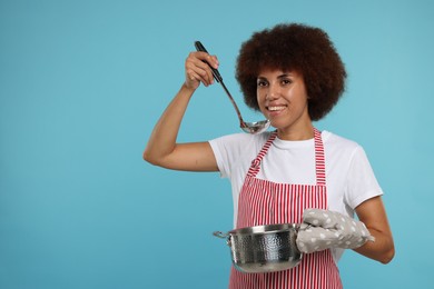 Photo of Happy young woman in apron holding ladle and cooking pot on light blue background. Space for text