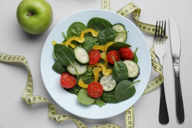 Photo of Measuring tape, salad, apple and cutlery on light background, flat lay