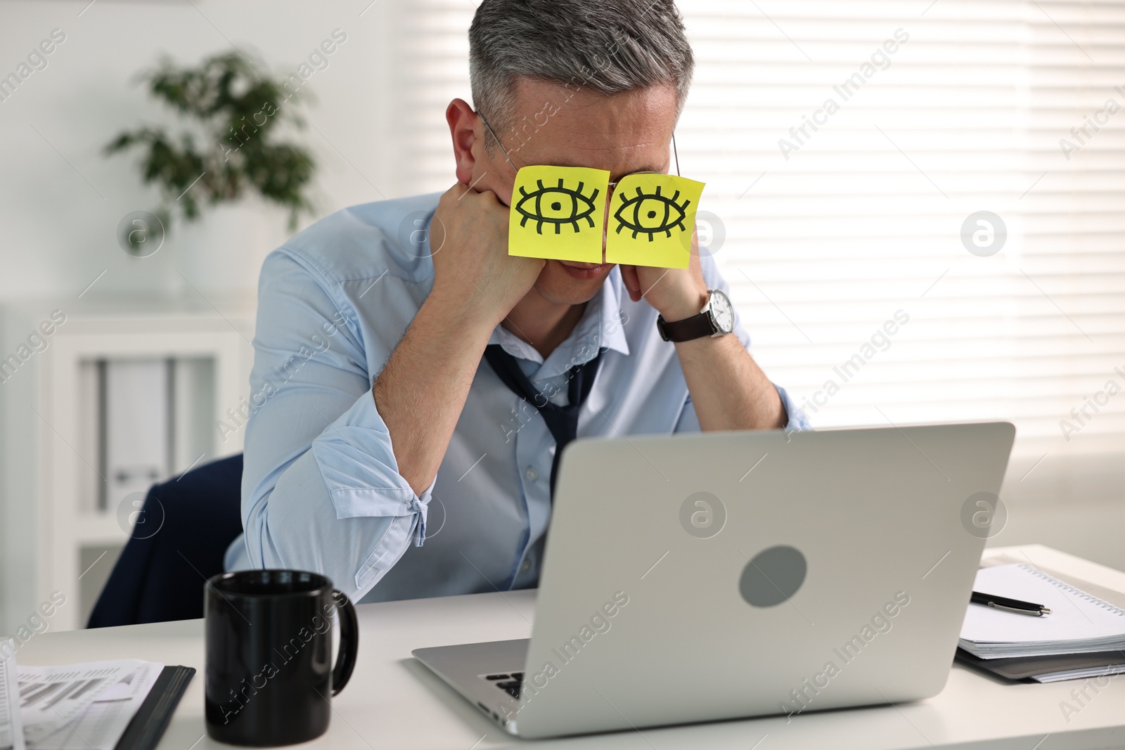 Photo of Man with fake eyes painted on sticky notes snoozing at workplace in office