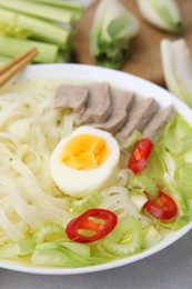 Photo of Bowl of delicious rice noodle soup with meat and egg on light grey table, closeup