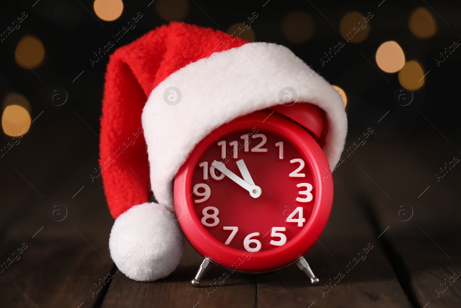 Photo of Alarm clock with decor on brown wooden table against blurred Christmas lights, closeup. New Year countdown