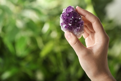 Woman holding beautiful purple amethyst gemstone on blurred green background, closeup. Space for text