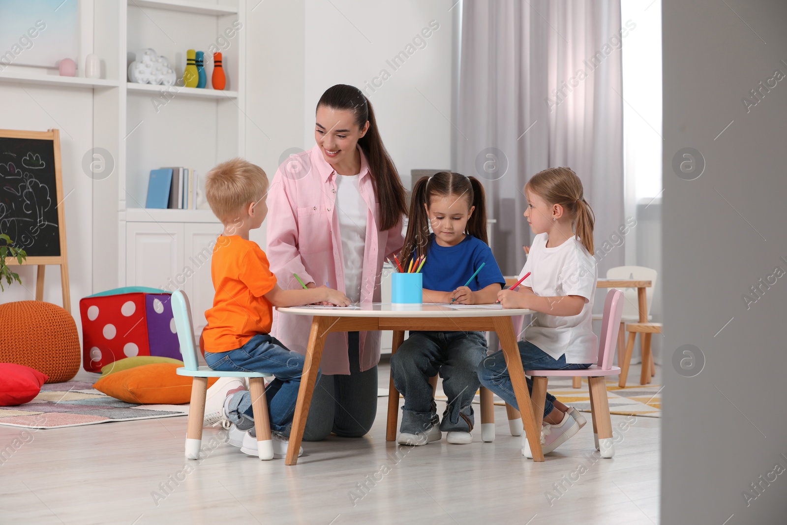 Photo of Nursery teacher and group of cute little children drawing at desk in kindergarten. Playtime activities