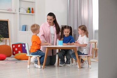Nursery teacher and group of cute little children drawing at desk in kindergarten. Playtime activities