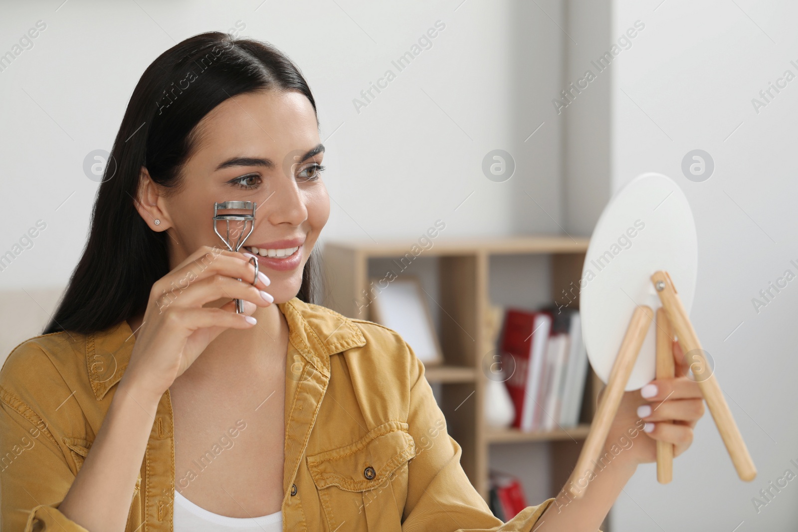 Photo of Beautiful young woman with mirror using eyelash curler indoors