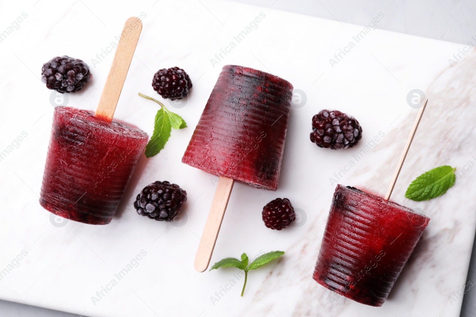 Photo of Tasty blackberry ice pops on white marble table, flat lay. Fruit popsicle