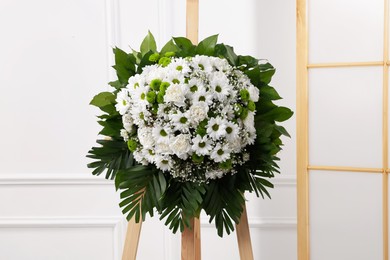 Funeral wreath of flowers on wooden stand near white wall
