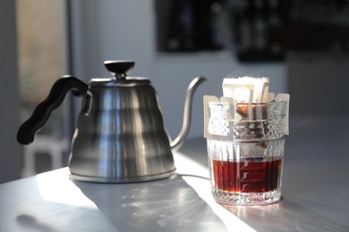 Photo of Glass with drip coffee bag and kettle on light grey table, closeup