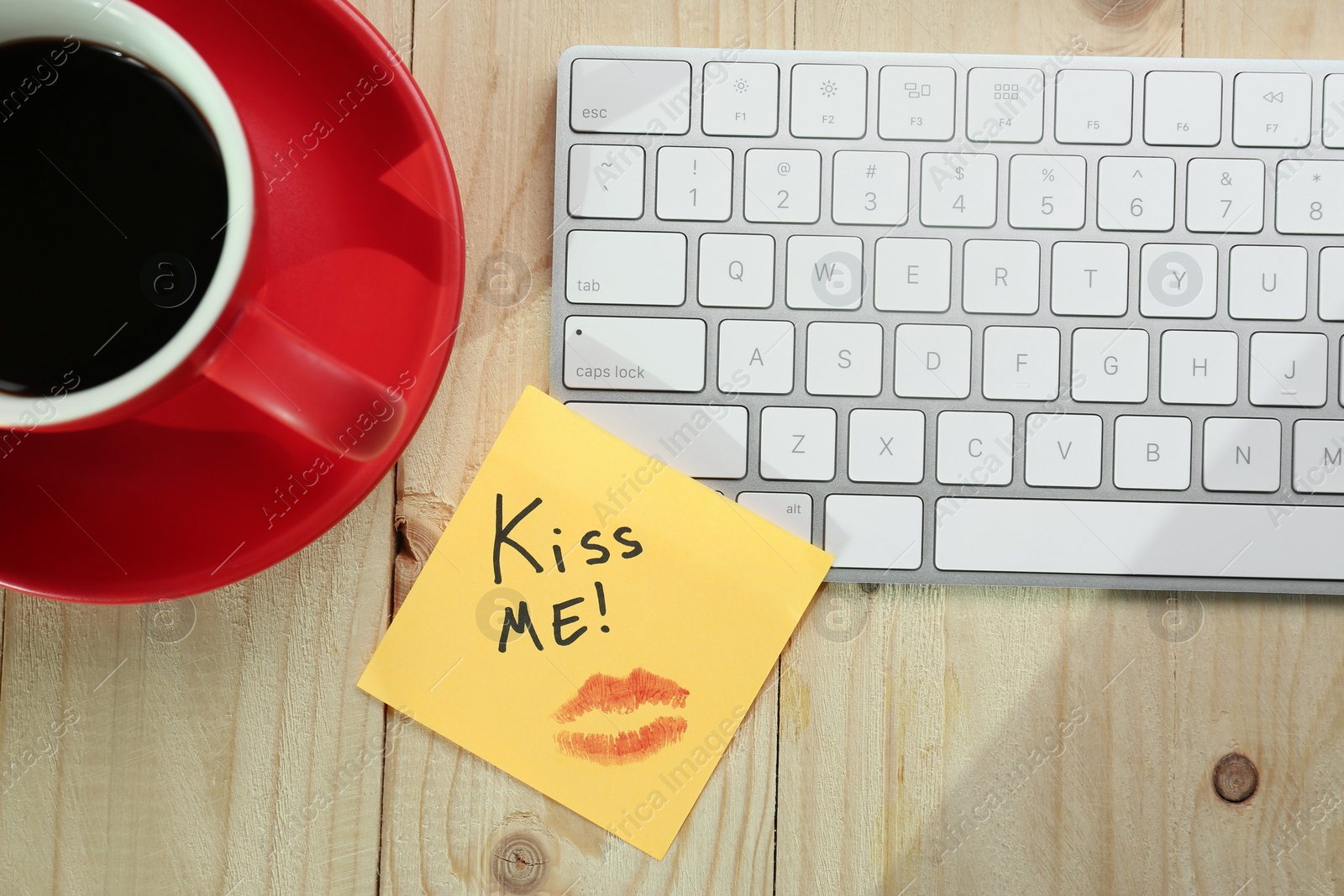 Photo of Sticky note with phrase Kiss Me, lipstick mark, cup of coffee and keyboard on wooden table, flat lay