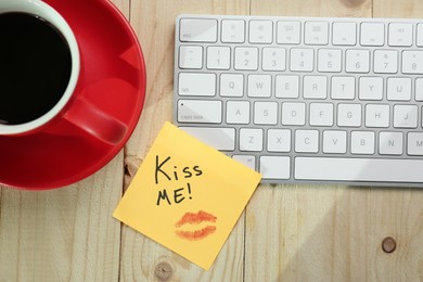 Photo of Sticky note with phrase Kiss Me, lipstick mark, cup of coffee and keyboard on wooden table, flat lay