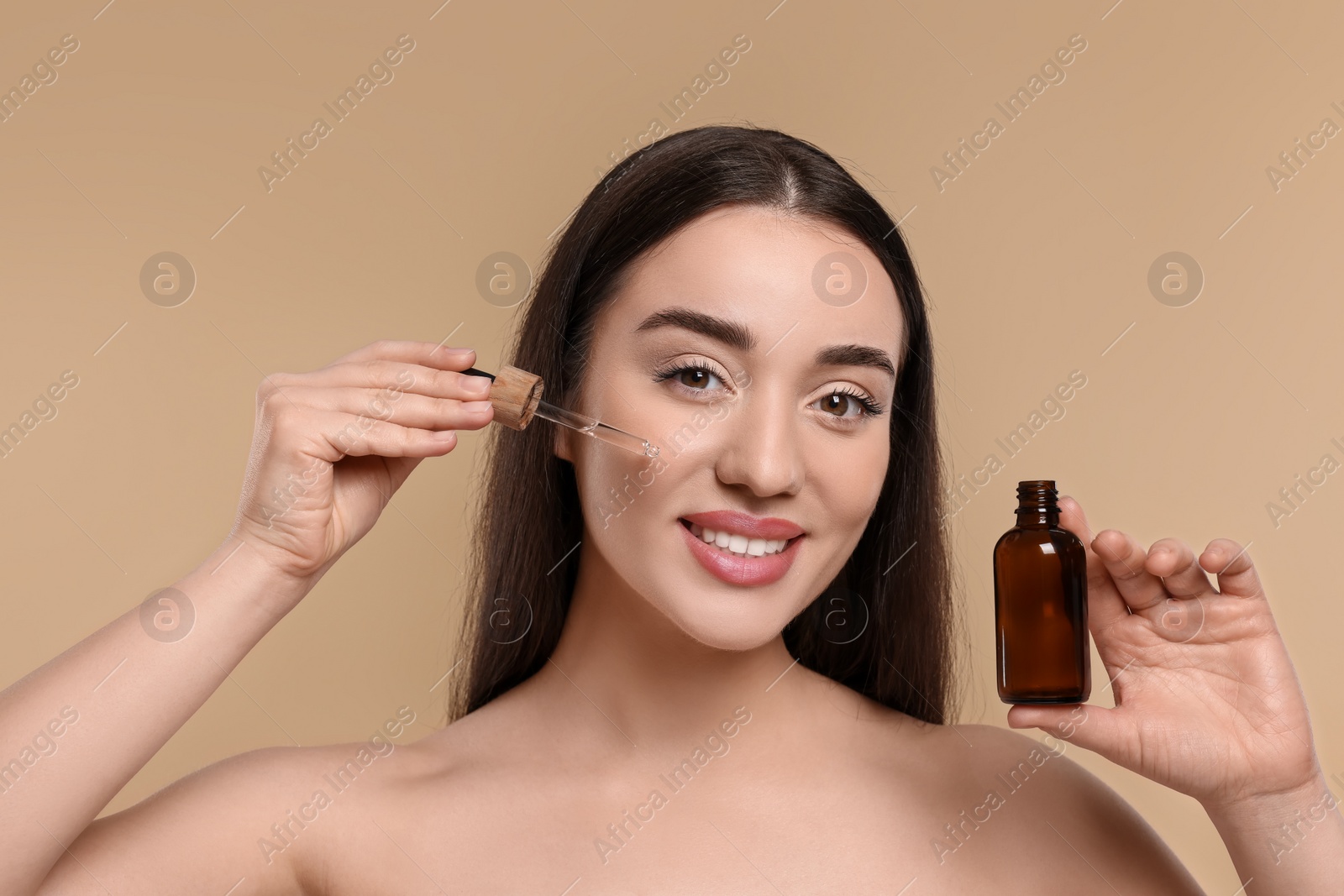 Photo of Happy young woman with bottle applying essential oil onto face on beige background