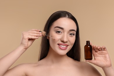 Happy young woman with bottle applying essential oil onto face on beige background