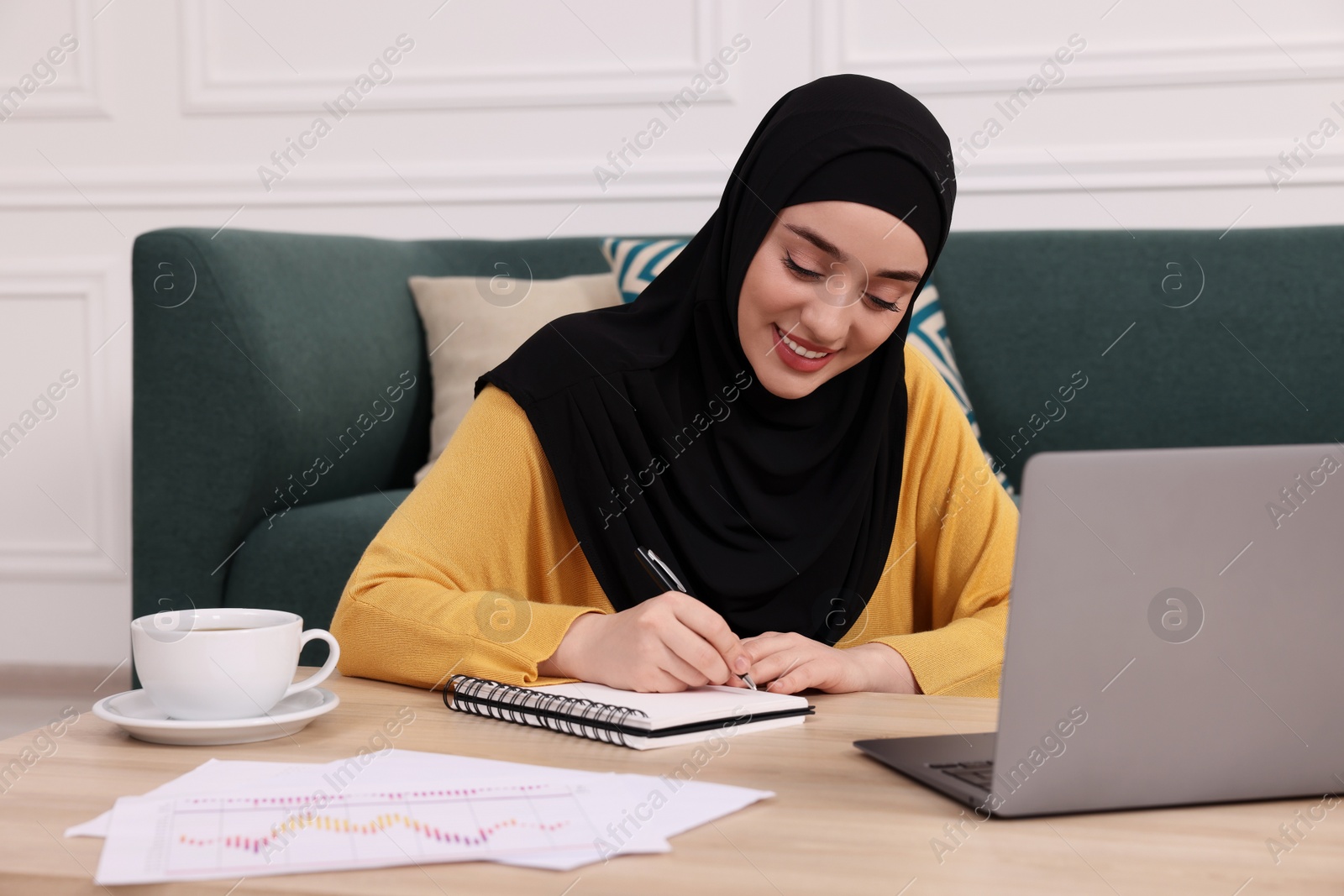 Photo of Muslim woman in hijab writing notes near laptop at wooden table indoors