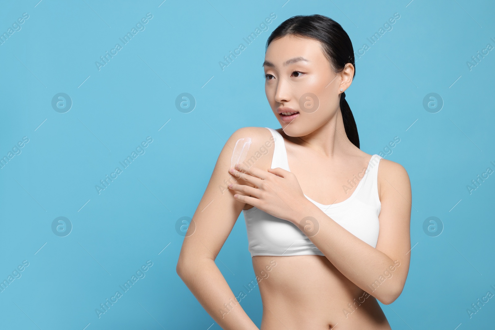 Photo of Beautiful young Asian woman applying body cream onto shoulder on light blue background, space for text