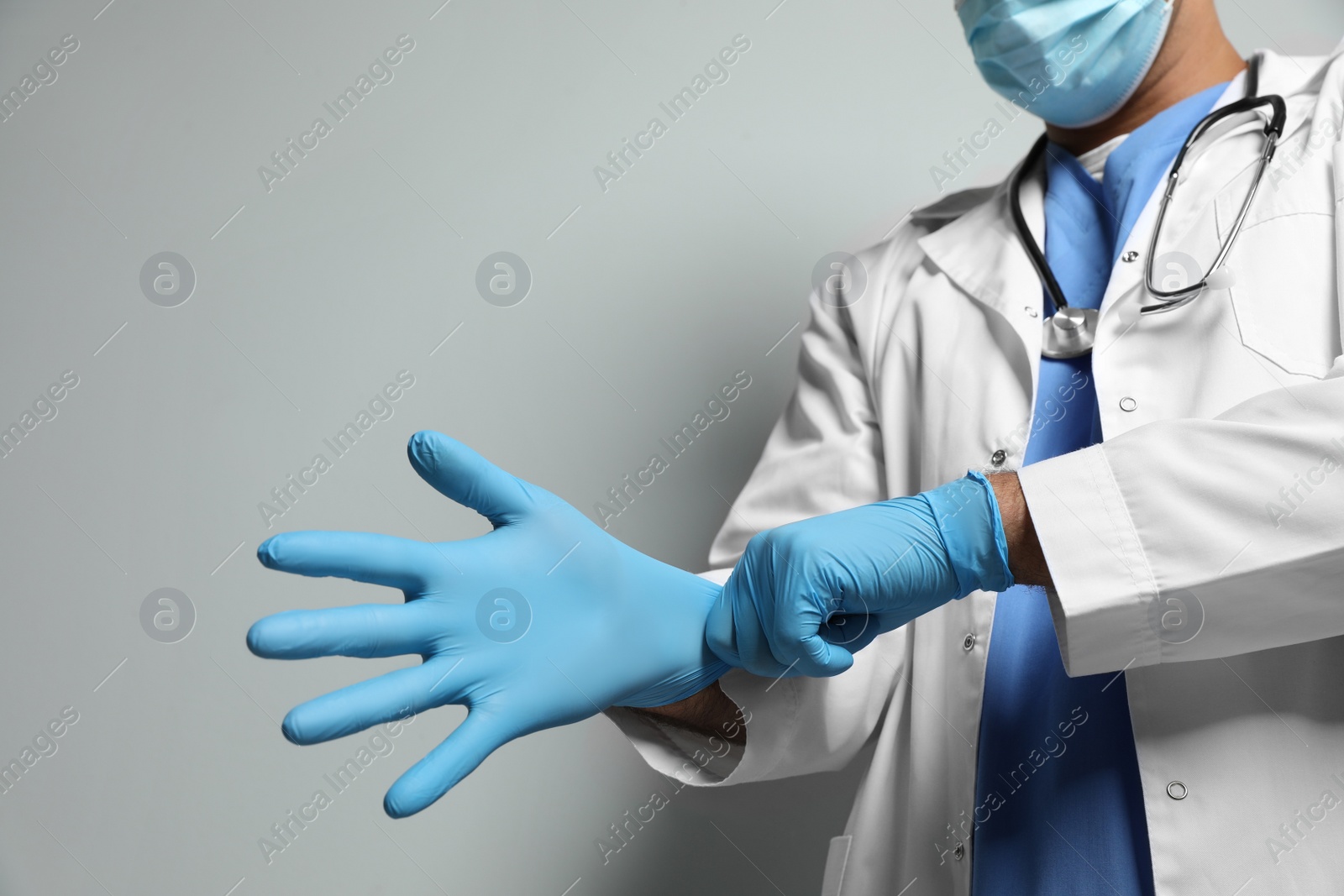 Photo of Doctor in protective mask putting on medical gloves against light grey background, closeup