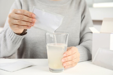 Woman pouring powder from medicine sachet into glass with water at table, closeup