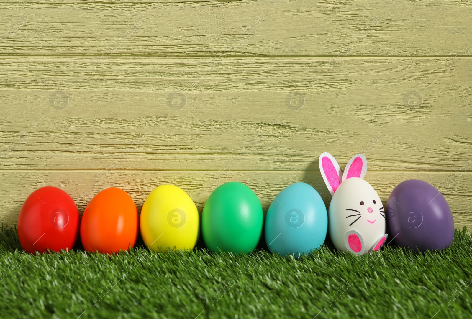 Photo of Bright eggs and white one as Easter bunny on green grass against wooden background