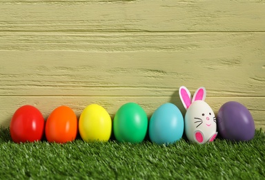 Bright eggs and white one as Easter bunny on green grass against wooden background