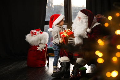 Photo of Santa Claus giving Christmas gift to little boy near window indoors