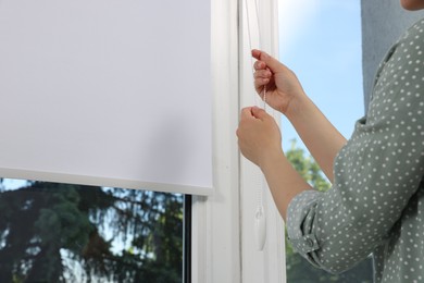 Woman opening white roller blind on window indoors, closeup