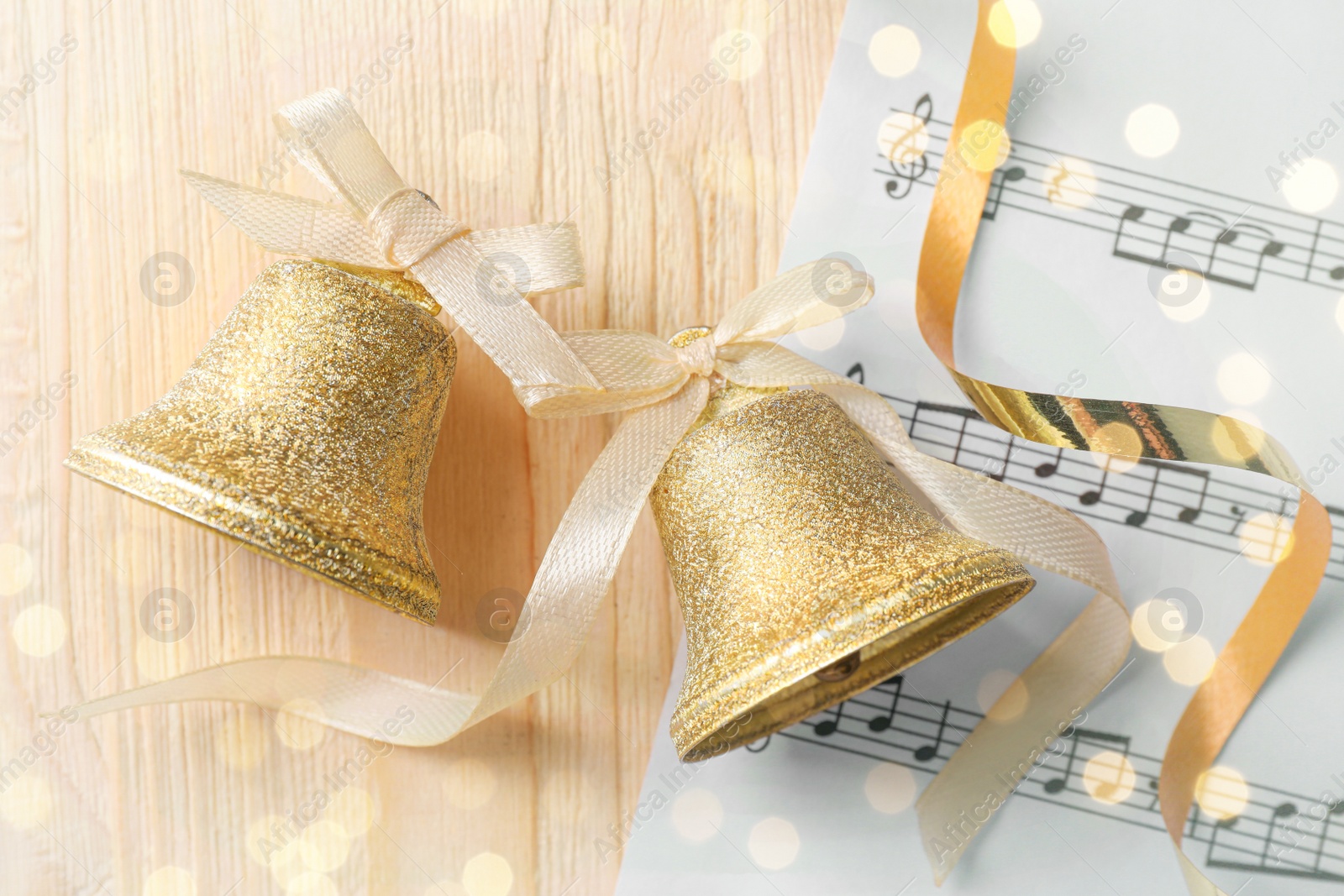 Photo of Golden shiny bells with white bows and music sheet on wooden table, flat lay. Christmas decoration