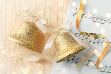 Photo of Golden shiny bells with white bows and music sheet on wooden table, flat lay. Christmas decoration