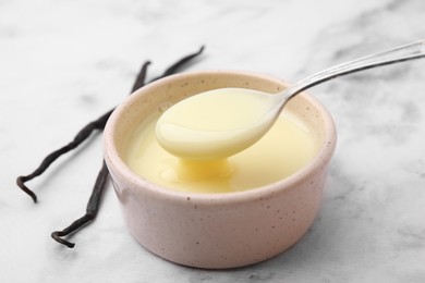Photo of Condensed milk flowing down from spoon into bowl and vanilla pods on white marble table, closeup