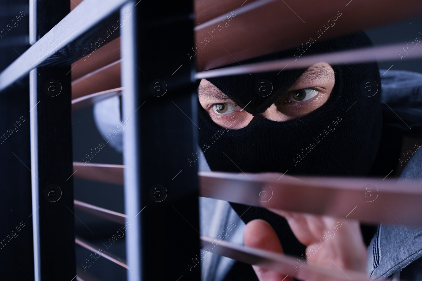 Photo of Masked man spying through window blinds indoors. Criminal offence