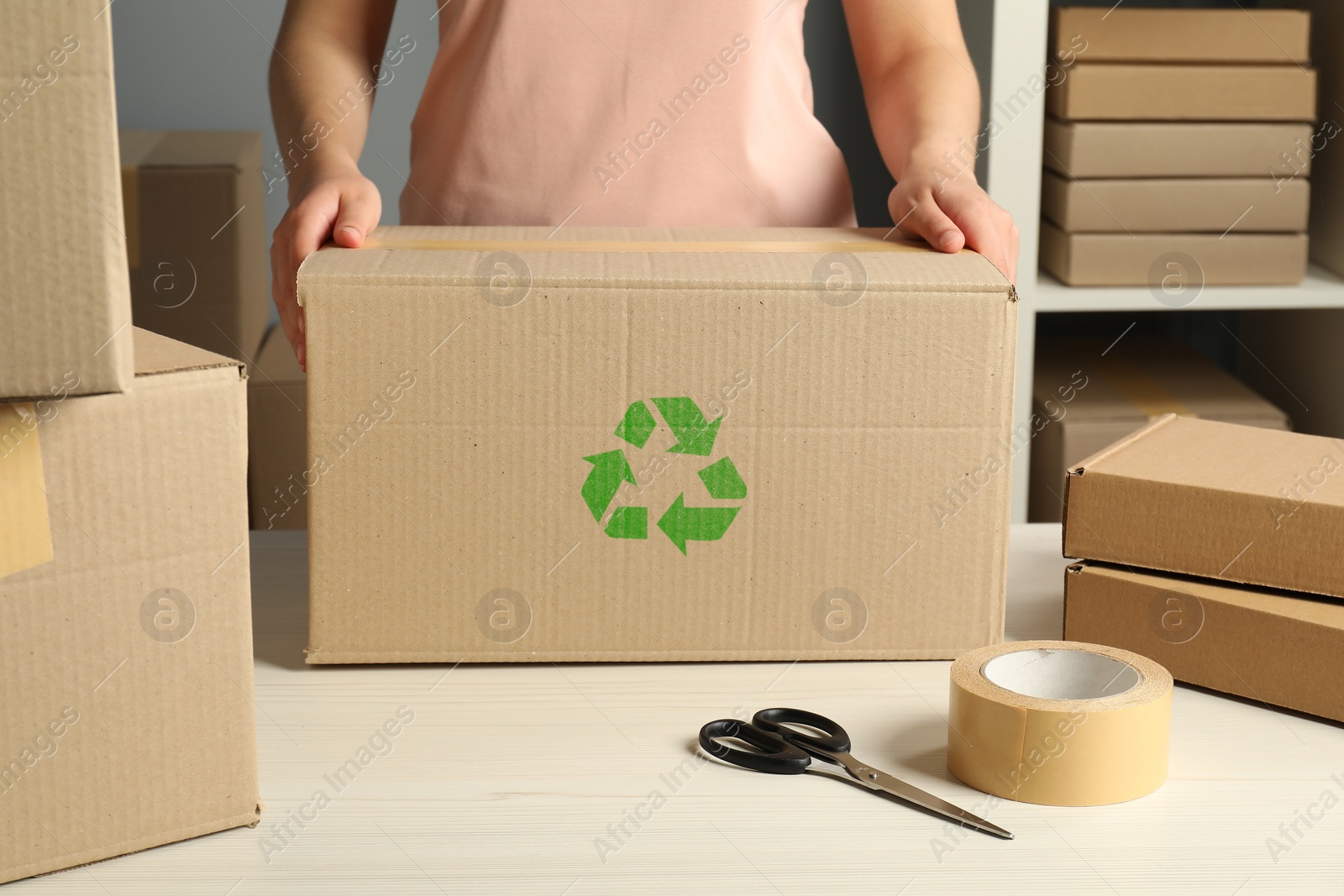 Image of Woman holding cardboard box with recycling sign stamp at white table indoors, closeup