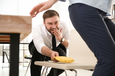 Young man putting whoopee cushion on chair while his colleague sitting down in office, closeup. Funny joke