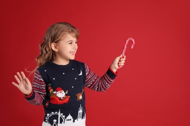 Photo of Cute little girl in Christmas sweater holding sweet candy cane against red background
