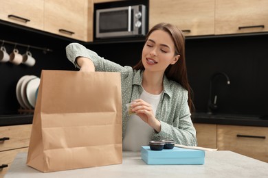 Photo of Beautiful young woman unpacking her order from sushi restaurant at table in kitchen