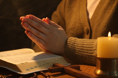 Woman praying at table with burning candle and Bible, closeup