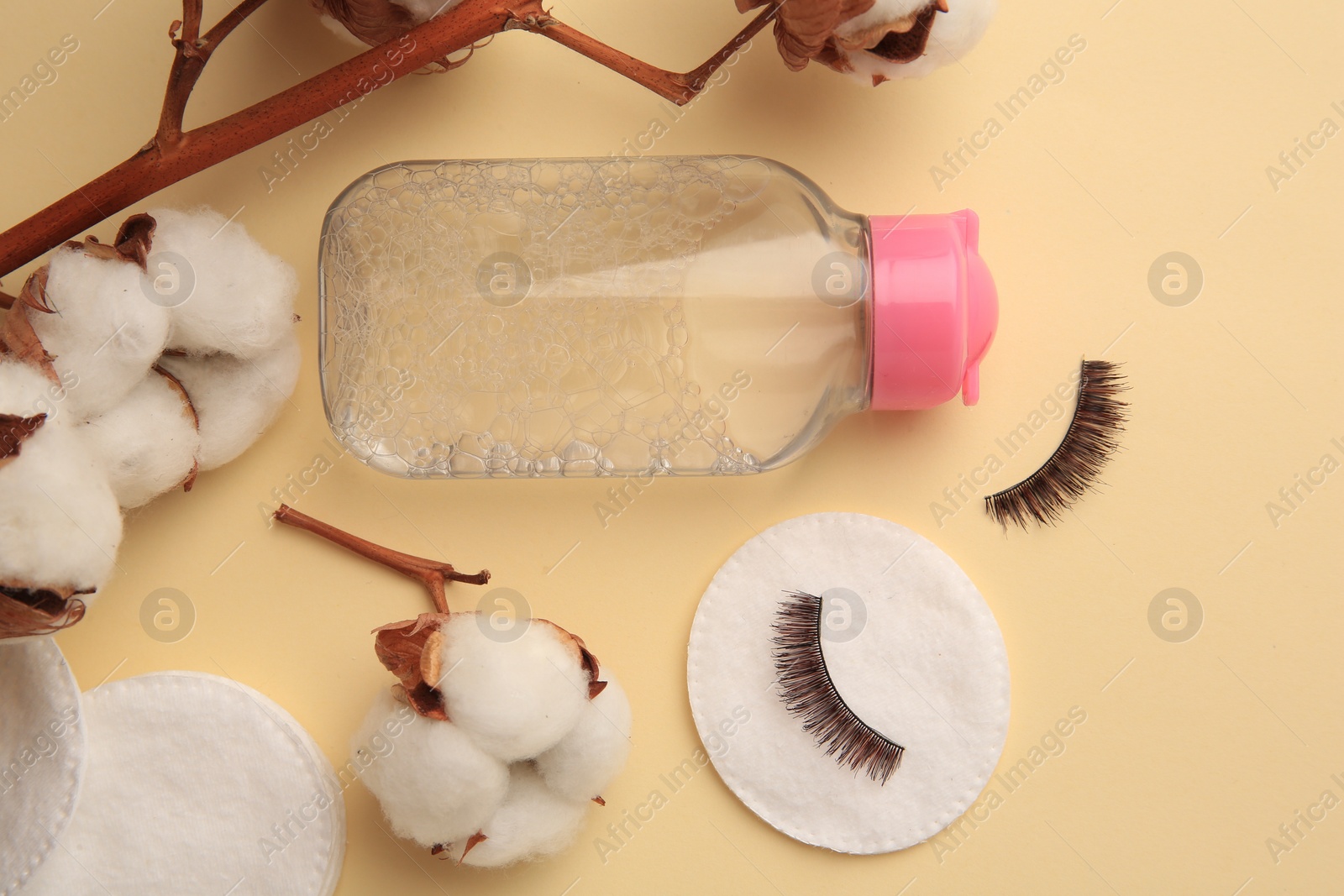 Photo of Bottle of makeup remover, cotton flowers, pads and false eyelashes on yellow background, flat lay