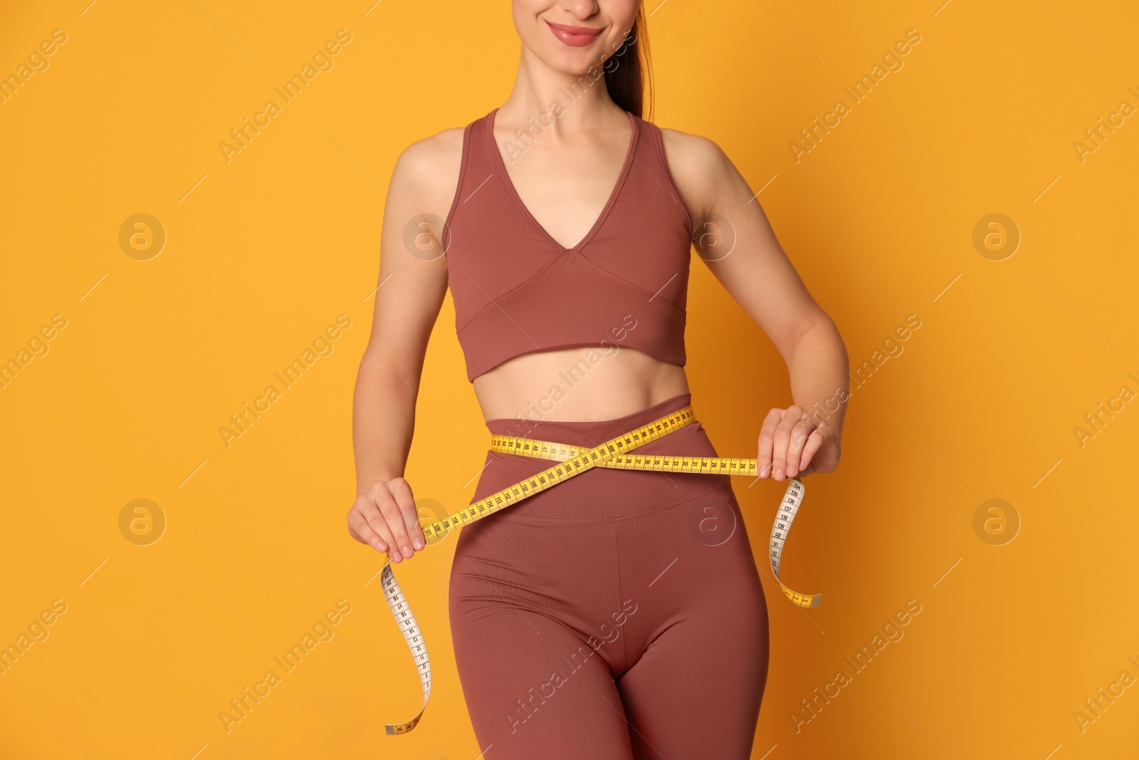 Photo of Happy woman in sportswear measuring waist with tape on yellow background, closeup