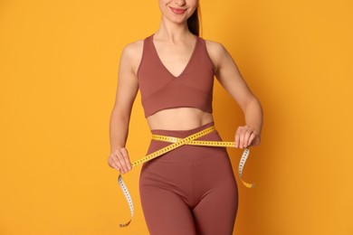 Happy woman in sportswear measuring waist with tape on yellow background, closeup