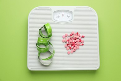 Photo of Scales with weight loss pills and measuring tape on green background, top view