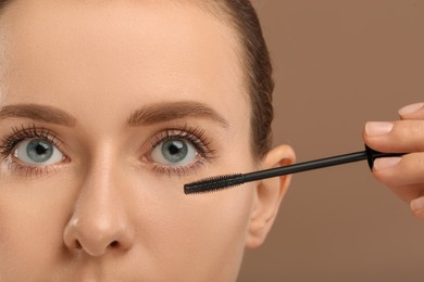 Woman applying mascara onto eyelashes against light brown background, closeup