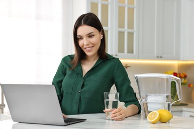 Photo of Woman with glass of water and filter jug near laptop in kitchen