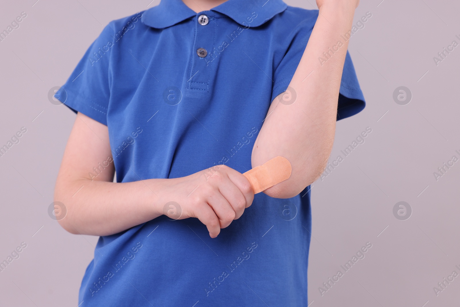 Photo of Little boy putting sticking plaster onto elbow against light grey background, closeup