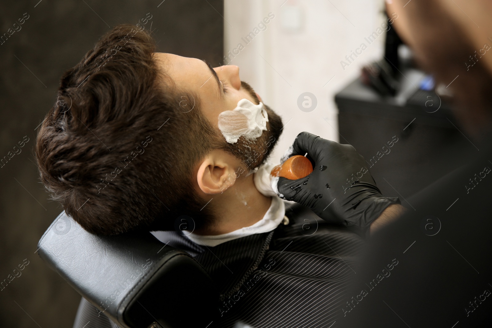 Photo of Professional hairdresser applying shaving foam onto client's skin in barbershop