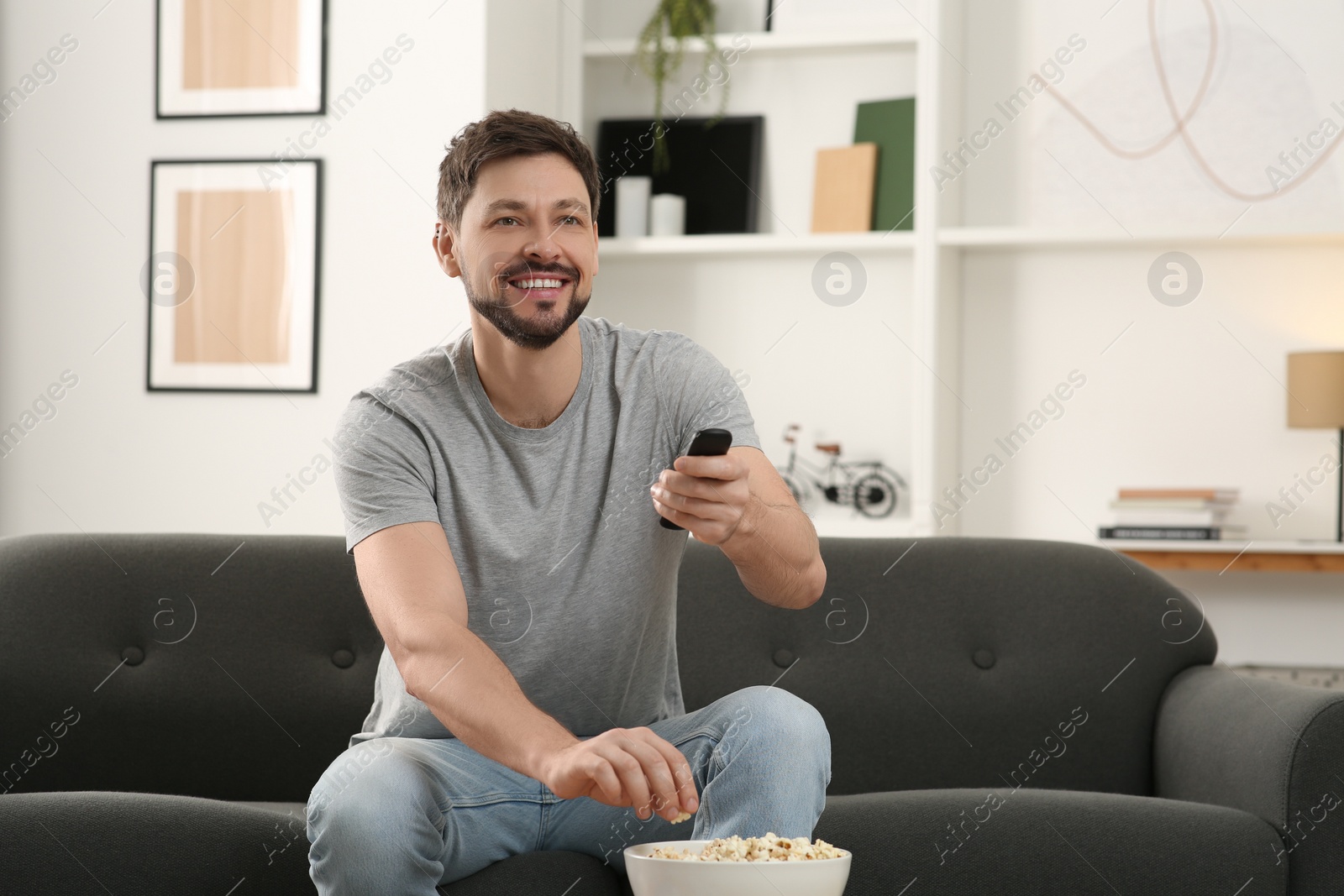 Photo of Happy man changing TV channels with remote control on sofa at home