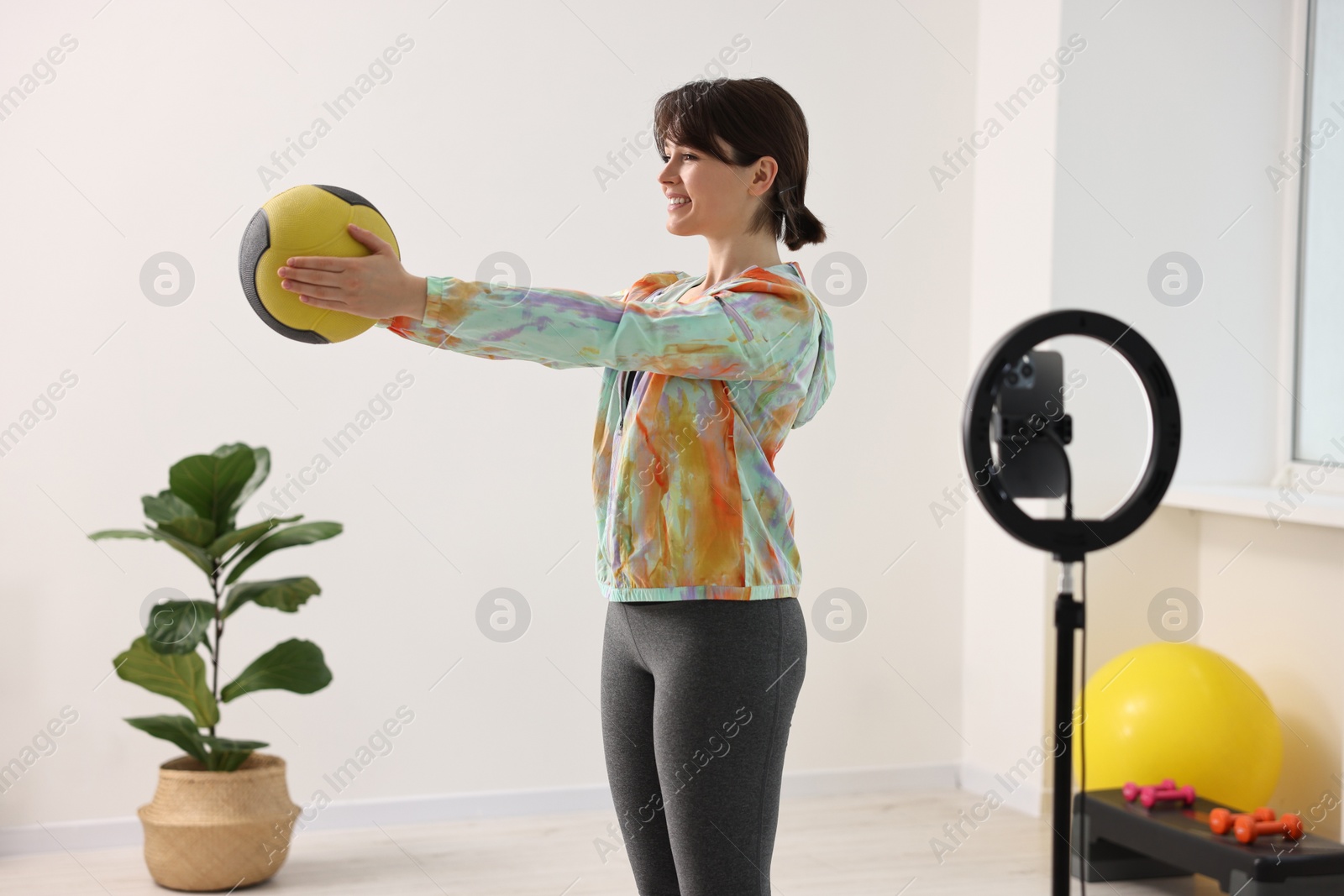 Photo of Happy sports blogger training with medicine ball while streaming online fitness lesson with smartphone at home