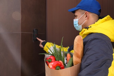 Photo of Courier in medical mask holding paper bag with groceries and ringing doorbell outdoors. Delivery service during quarantine due to Covid-19 outbreak