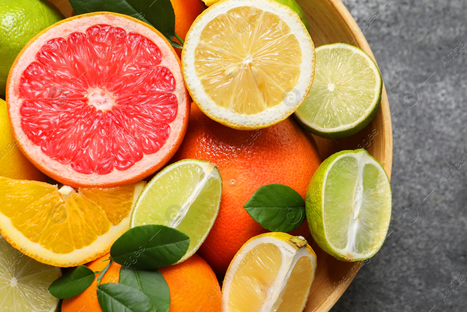 Photo of Different fresh citrus fruits and leaves in bowl on grey textured table, top view