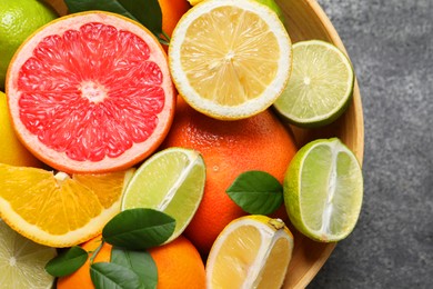 Photo of Different fresh citrus fruits and leaves in bowl on grey textured table, top view