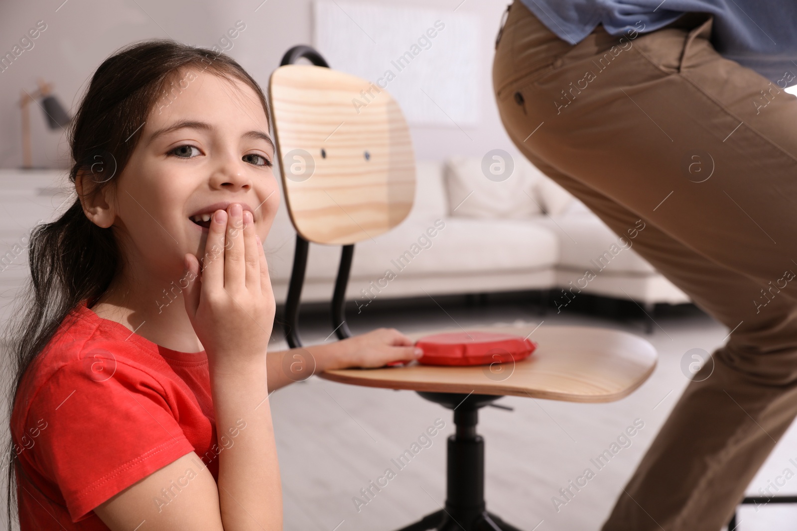 Photo of Cute little girl putting whoopee cushion on father's chair while he sitting down at home, closeup