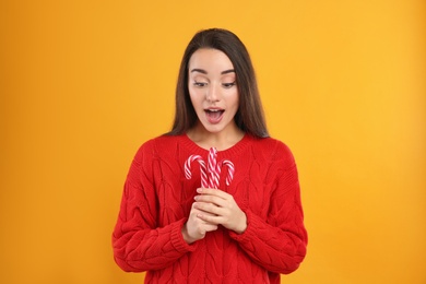 Photo of Surprised young woman in red sweater holding candy canes on yellow background. Celebrating Christmas